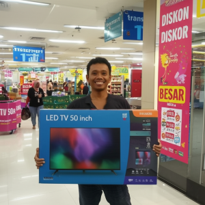 Seorang pria tersenyum sambil memegang kotak LED TV 50 inch di depan toko Transmart yang ramai dengan dekorasi diskon.