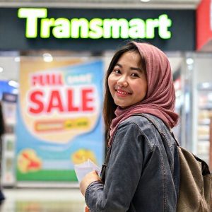 Seorang wanita sedang tersenyum sambil memegang kantong belanja Transmart, di belakangnya terlihat banner promosi 